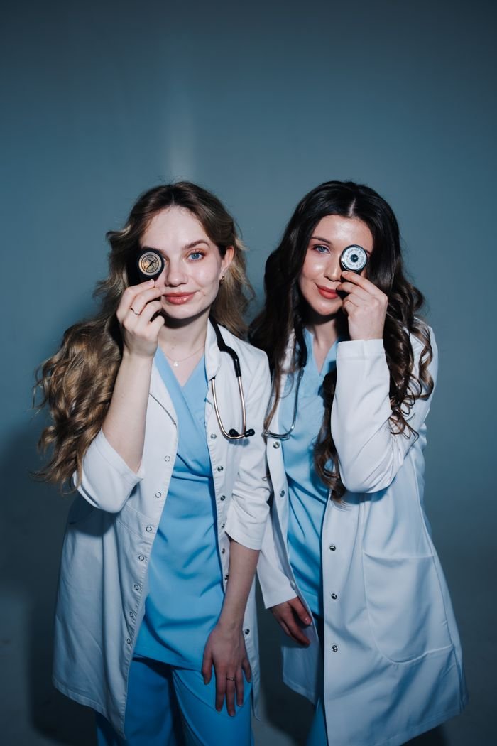 Two female doctors in lab coats, playfully covering their eyes with medical tools, highlighting fun in healthcare.
