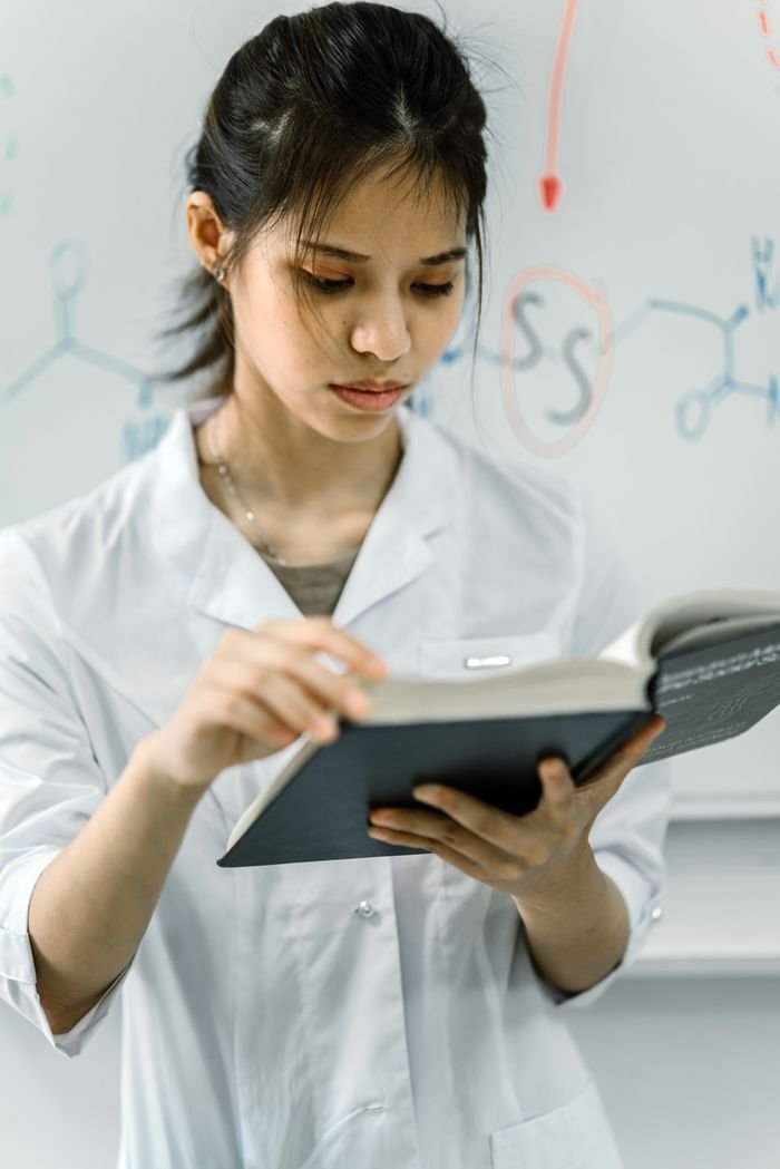 Scientist in lab coat reading a book in front of a chemistry-themed whiteboard.